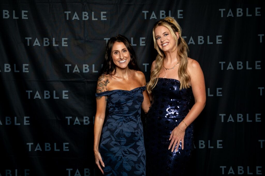 Two women in formal dresses pose in front of a black backdrop with "TABLE" repeated
