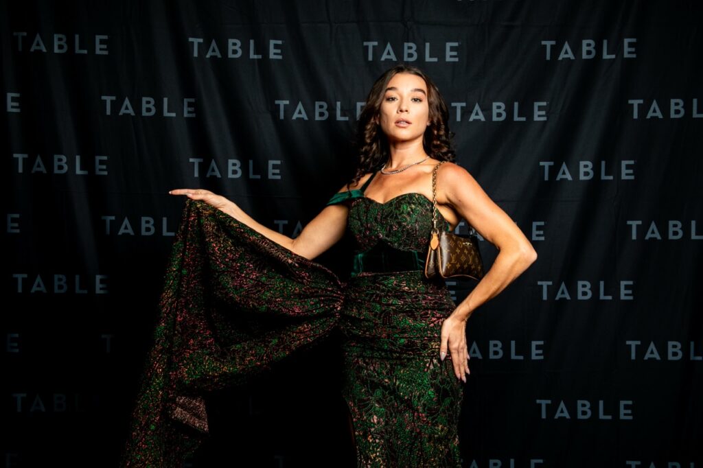 Woman in a green sequined gown poses in front of a black backdrop with "TABLE" logo.