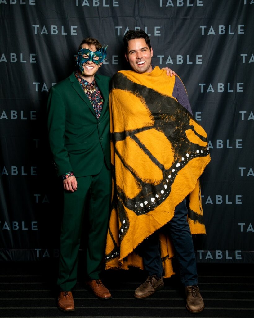 Two men pose for a photo at a TABLE event, one in a green suit and mask, the other draped in a monarch butterfly wing costume.
