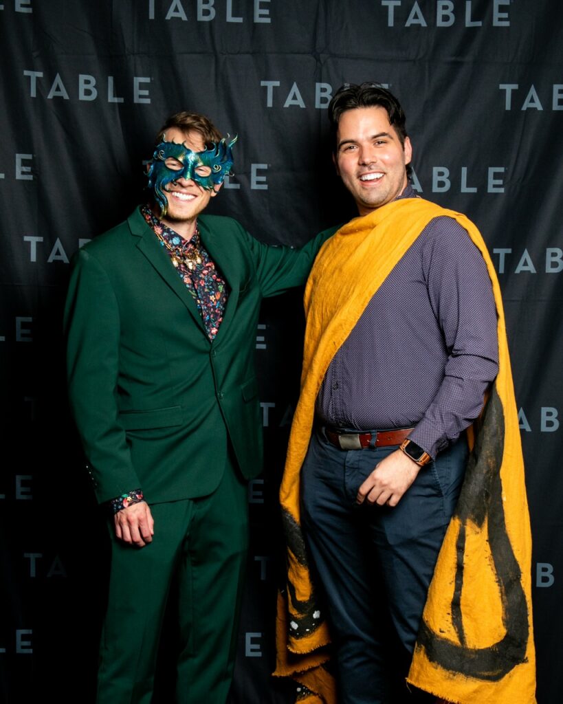 Two men in costumes pose for a photo at an event with a "TABLE" backdrop.