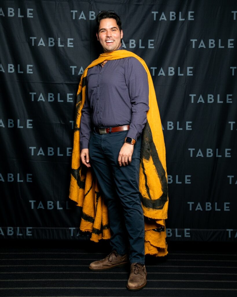 Man in a yellow butterfly-wing shawl smiles in front of a black TABLE backdrop.