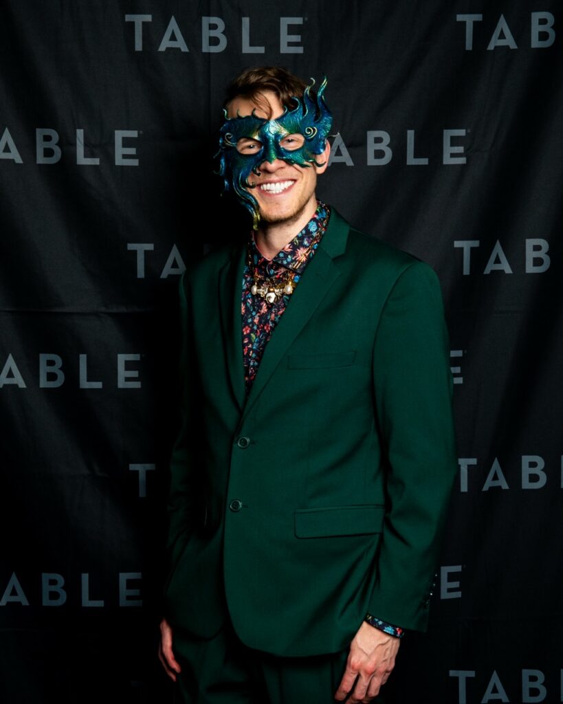Man in green suit and ornate mask smiles in front of a black backdrop with "TABLE" repeated.