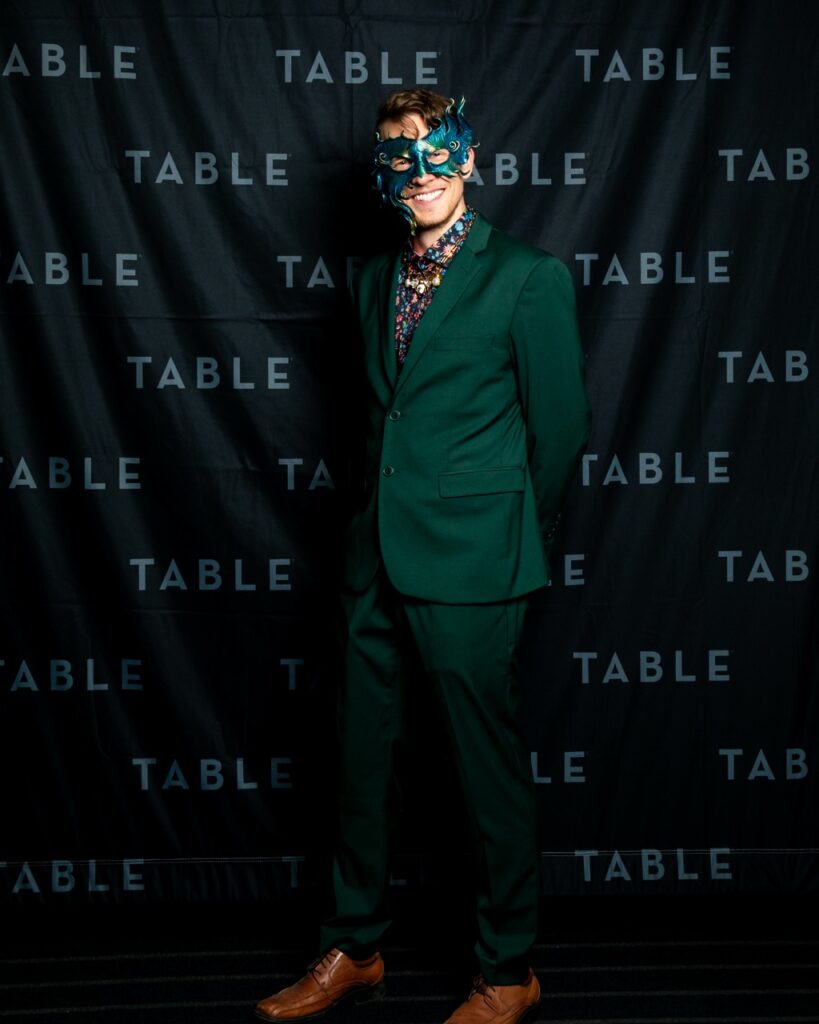 Man in green suit and ornate mask smiles in front of TABLE backdrop