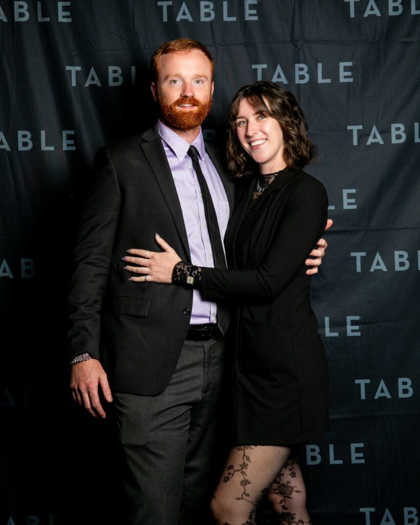 Couple posing in front of a black backdrop with "TABLE" logo at an event.