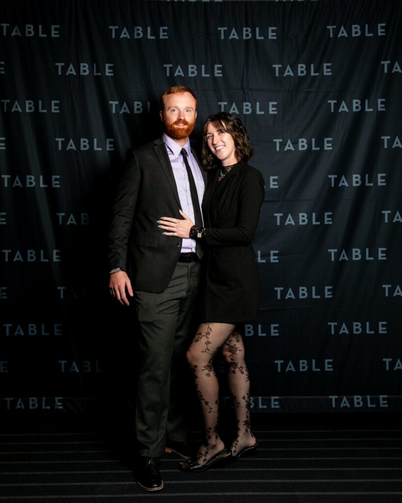 Man in suit and woman in black dress pose in front of a black backdrop with "TABLE" repeated