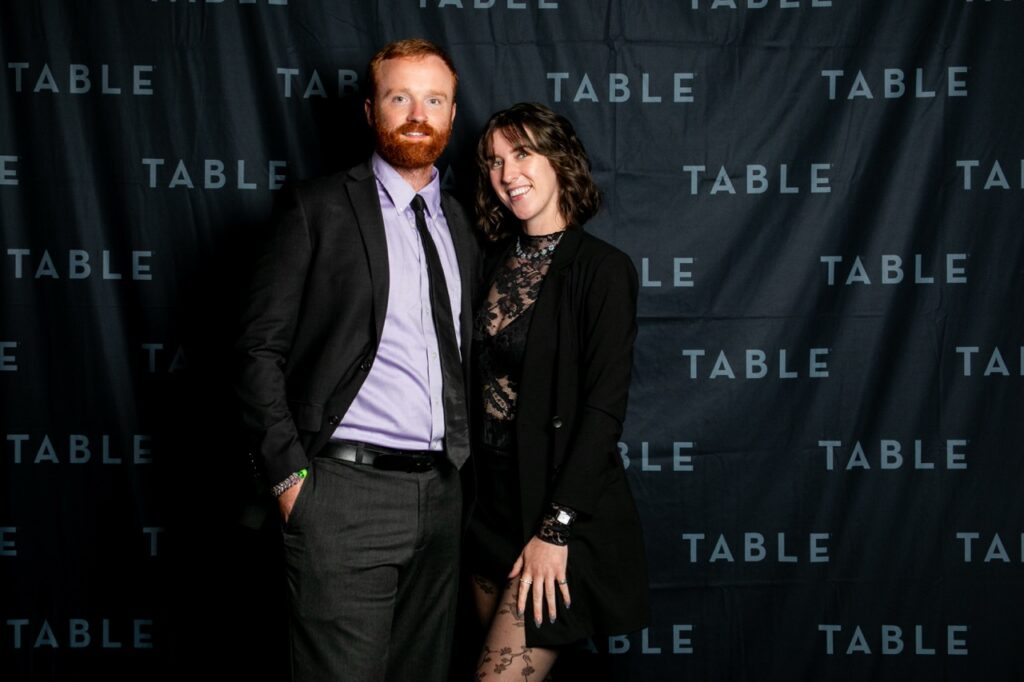 Couple posing for a photo in front of a black backdrop with "TABLE" printed repeatedly.