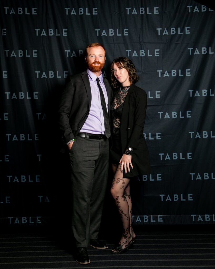 Couple poses in front of a black backdrop with "TABLE" repeated
