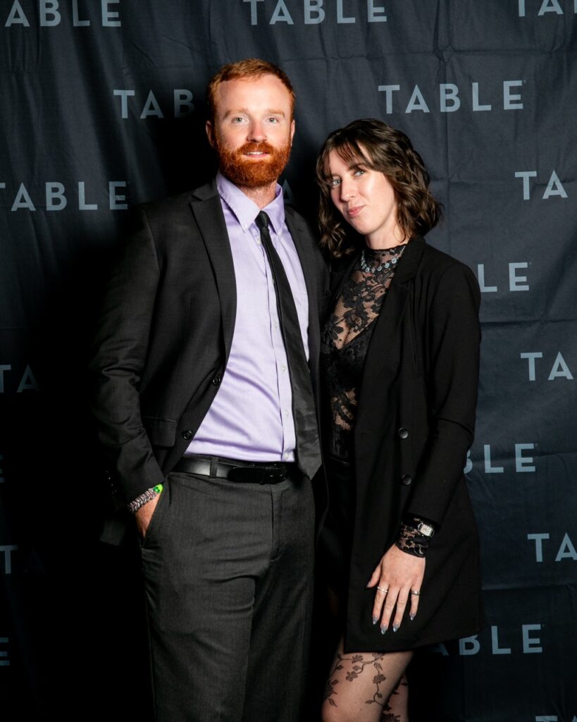 Couple in formal attire poses for a photo in front of a black backdrop with "TABLE" logo