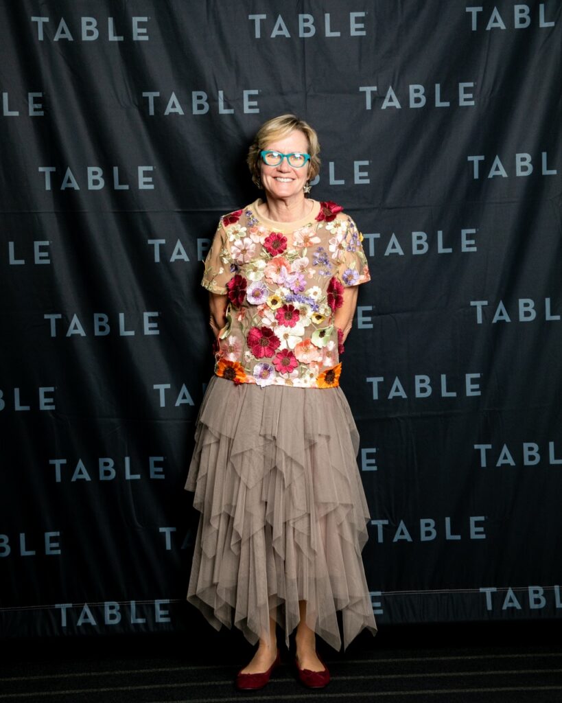 Woman in floral top and tiered skirt poses in front of a "TABLE" backdrop