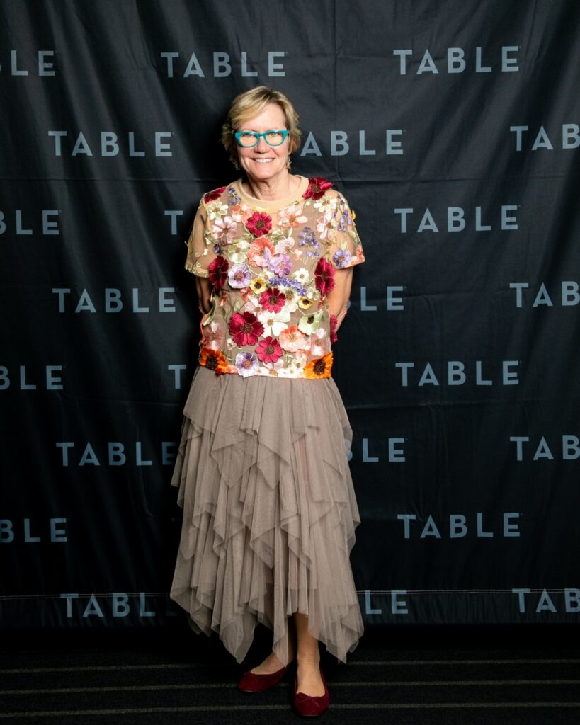 Woman in floral top and taupe tiered skirt smiles in front of a black "TABLE" backdrop.