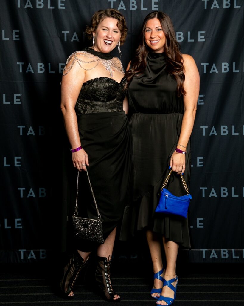 Two women in black dresses pose for a photo at an event with a black backdrop.