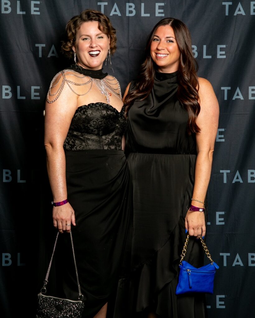 Two women in black dresses smile at the camera at an event with a "TABLE" backdrop.