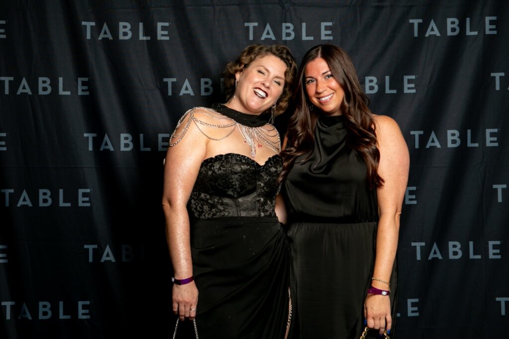 Two women in black dresses posing for a photo at an event with a "TABLE" backdrop.