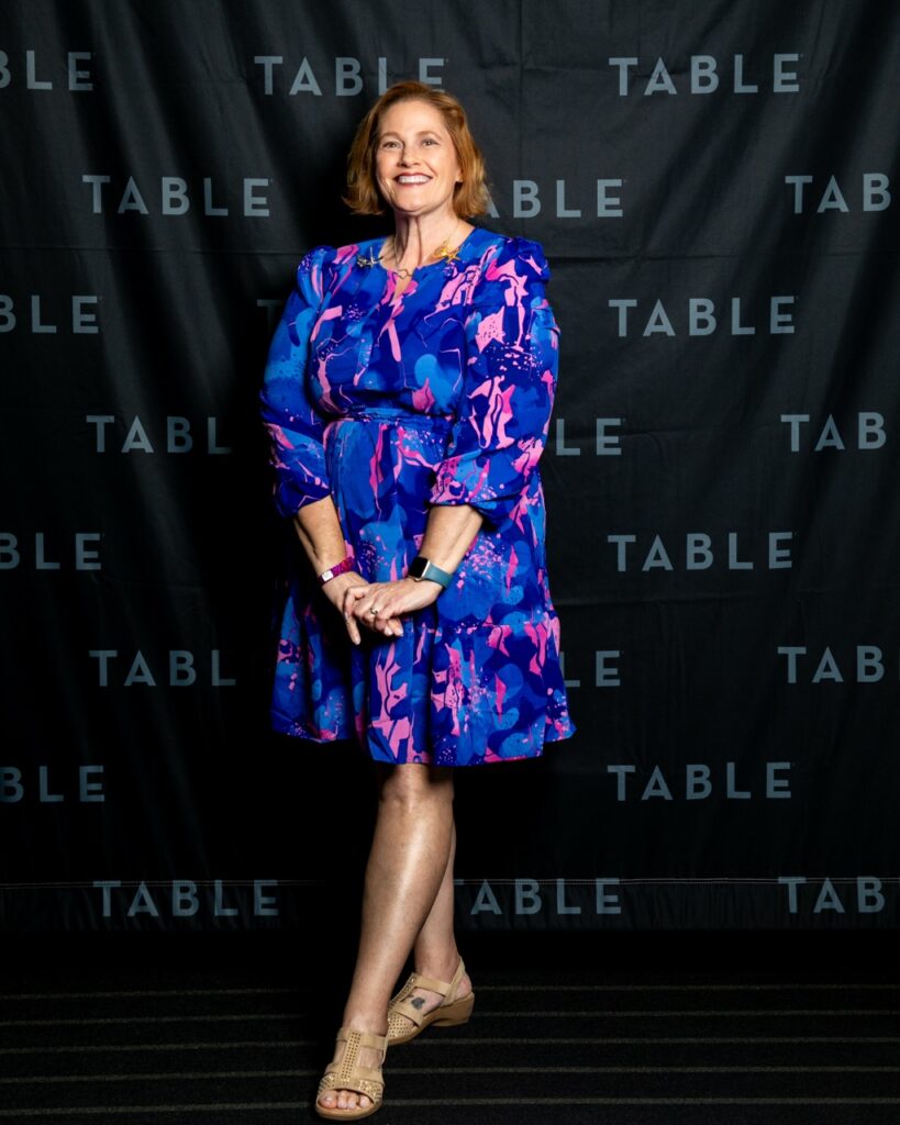Woman in a blue and pink floral dress smiles in front of a black backdrop with "TABLE" repeated.