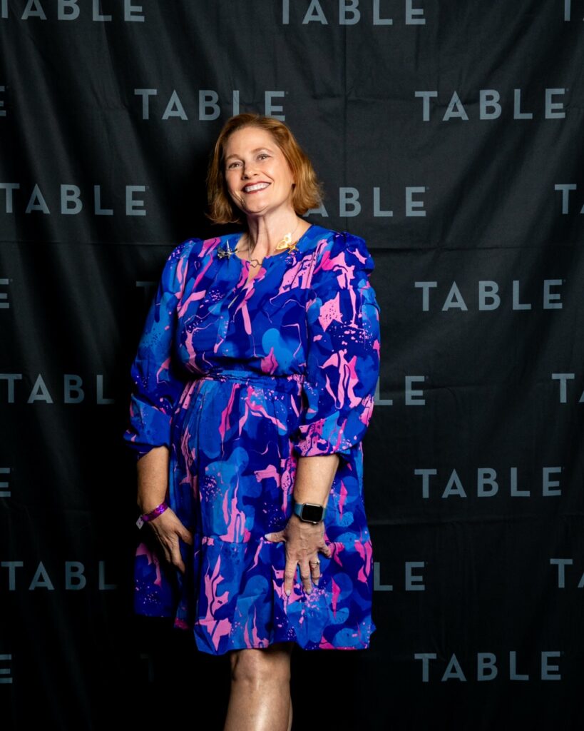 Woman in a blue and pink floral dress smiles at the camera in front of a black backdrop with "TABLE" repeated.