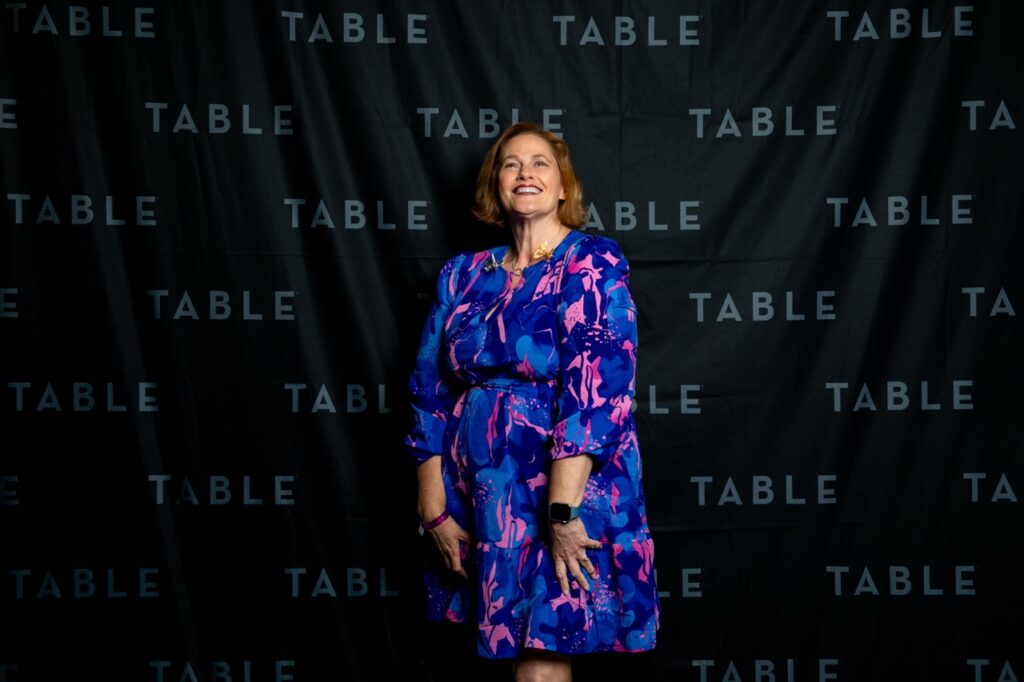 Woman in a blue and pink dress smiles in front of a black backdrop with repeating "TABLE" text.