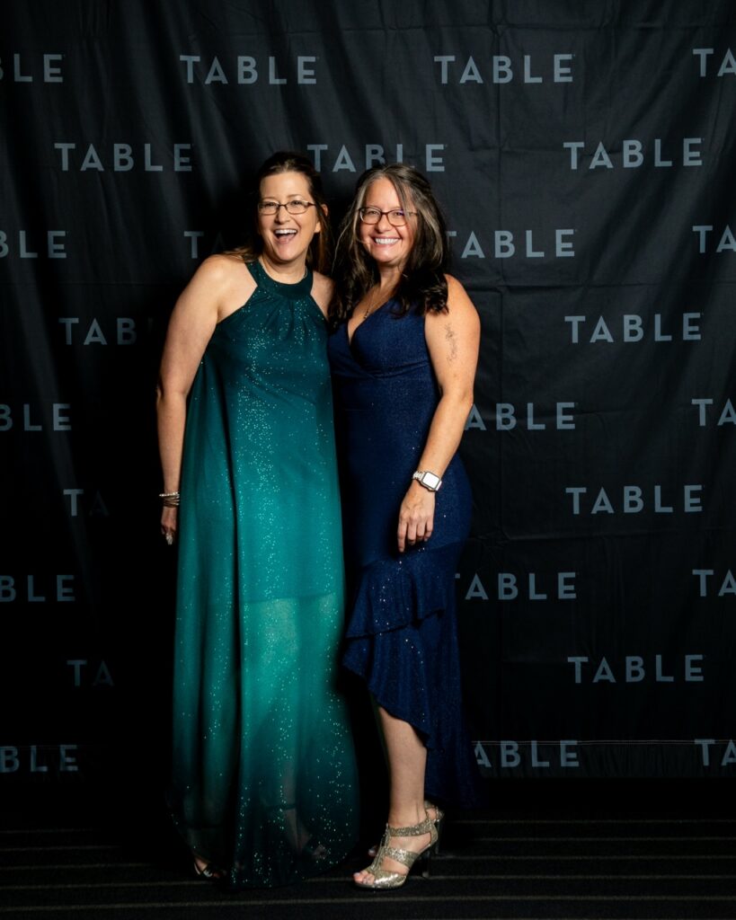 Two women in formal dresses pose for a photo in front of a backdrop with the word "TABLE" repeated.