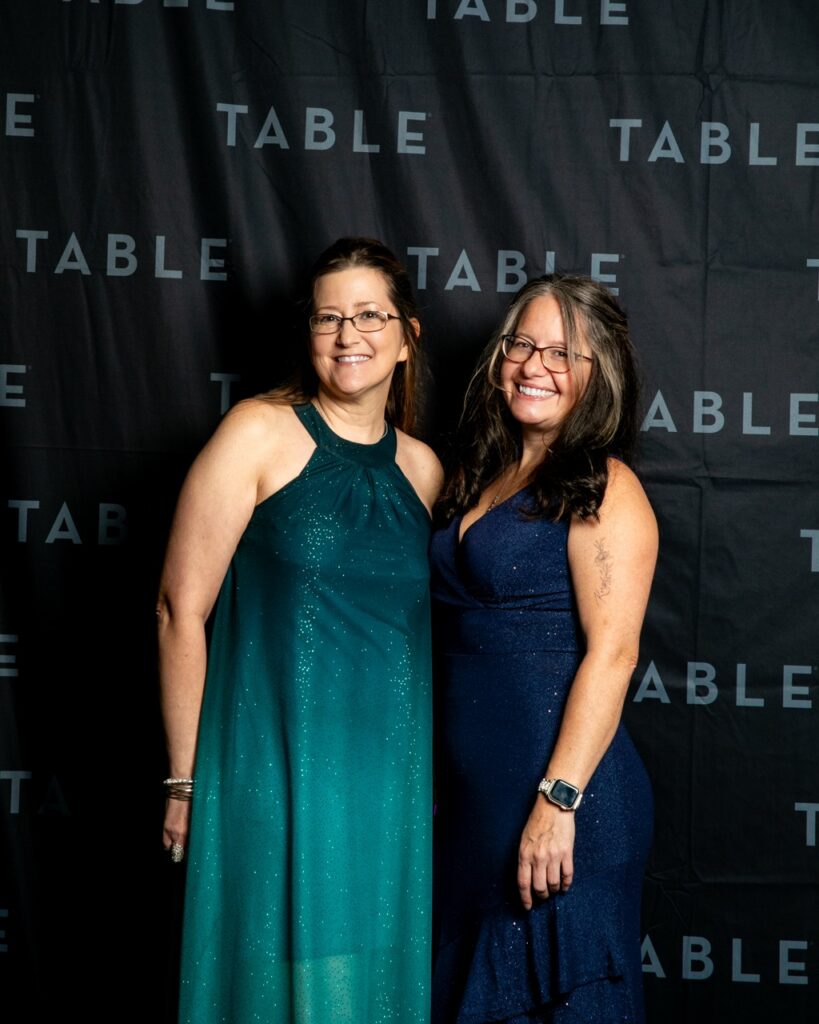 Two smiling women in formal dresses pose in front of a black backdrop with "TABLE" repeating.