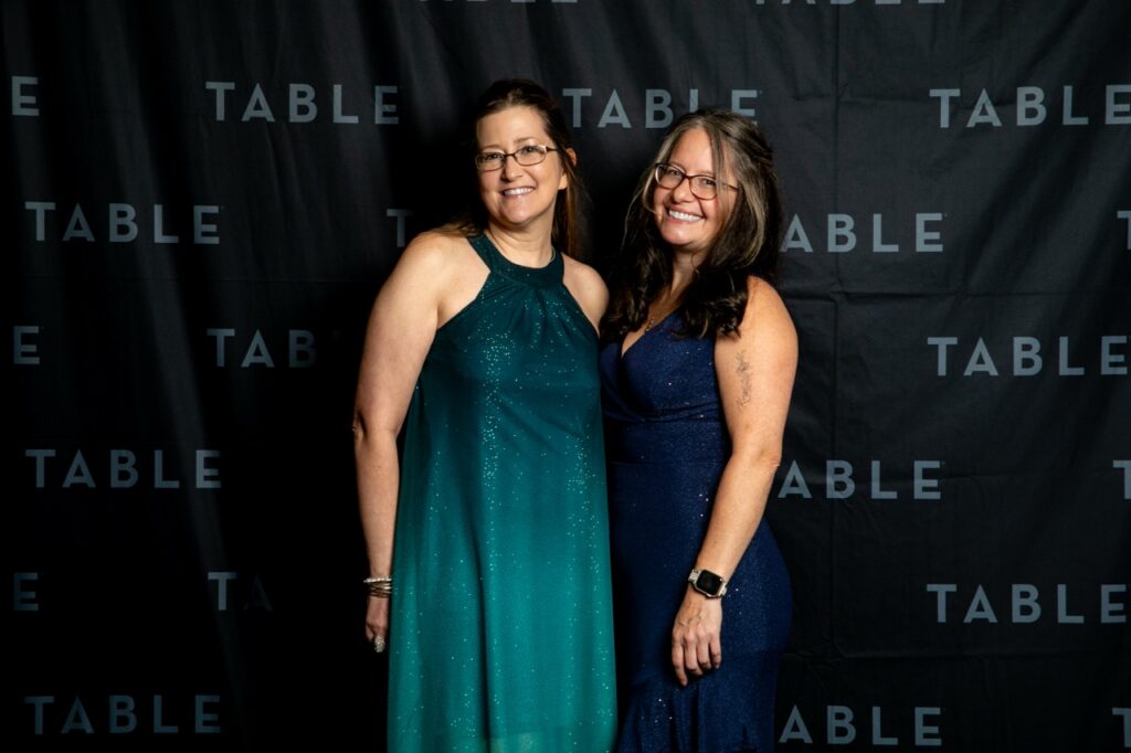 Two smiling women in formal dresses pose in front of a black backdrop with "TABLE" repeated.