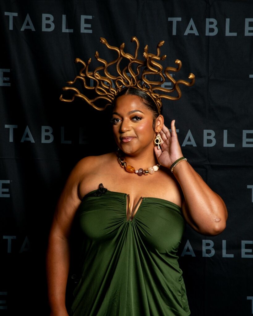 Woman in green dress and Medusa-inspired snake crown poses against a backdrop with "TABLE" text.