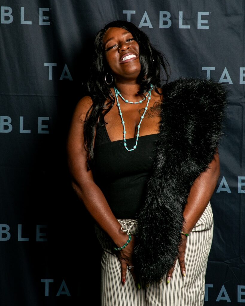 Woman in black tank top, striped pants, and faux fur stole smiles at camera.