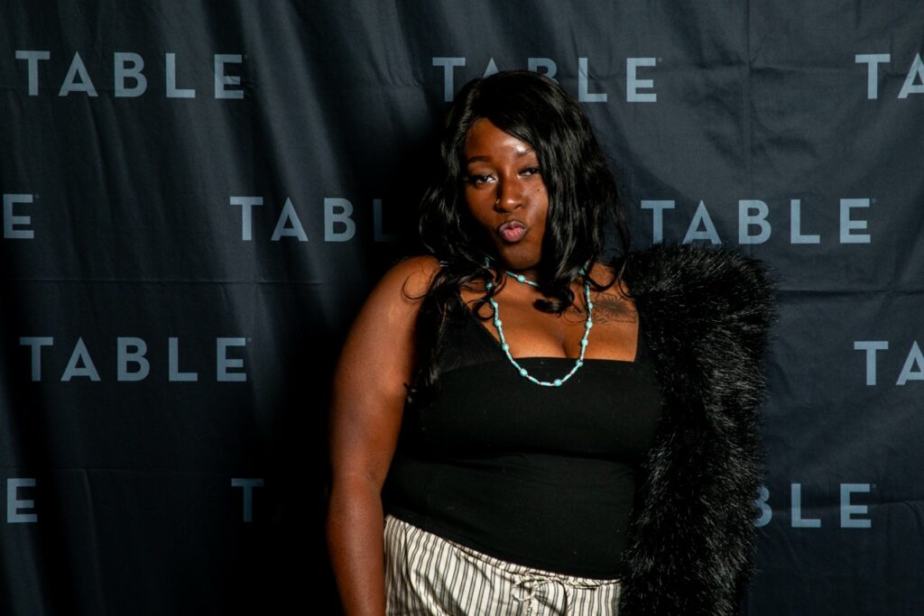 Woman in black top and striped pants poses in front of a black backdrop with "TABLE" logo.