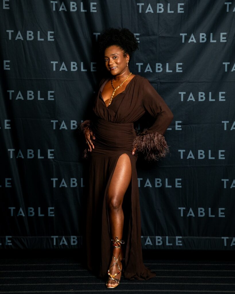 Woman in a brown dress with a high slit and feathered sleeves poses in front of a backdrop with "TABLE" repeated.