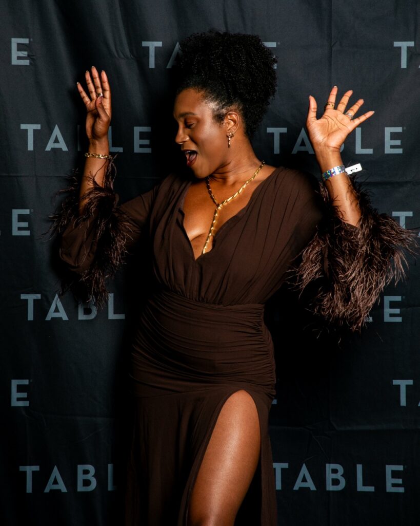 Woman in a brown dress with feathered sleeves posing with her hands up