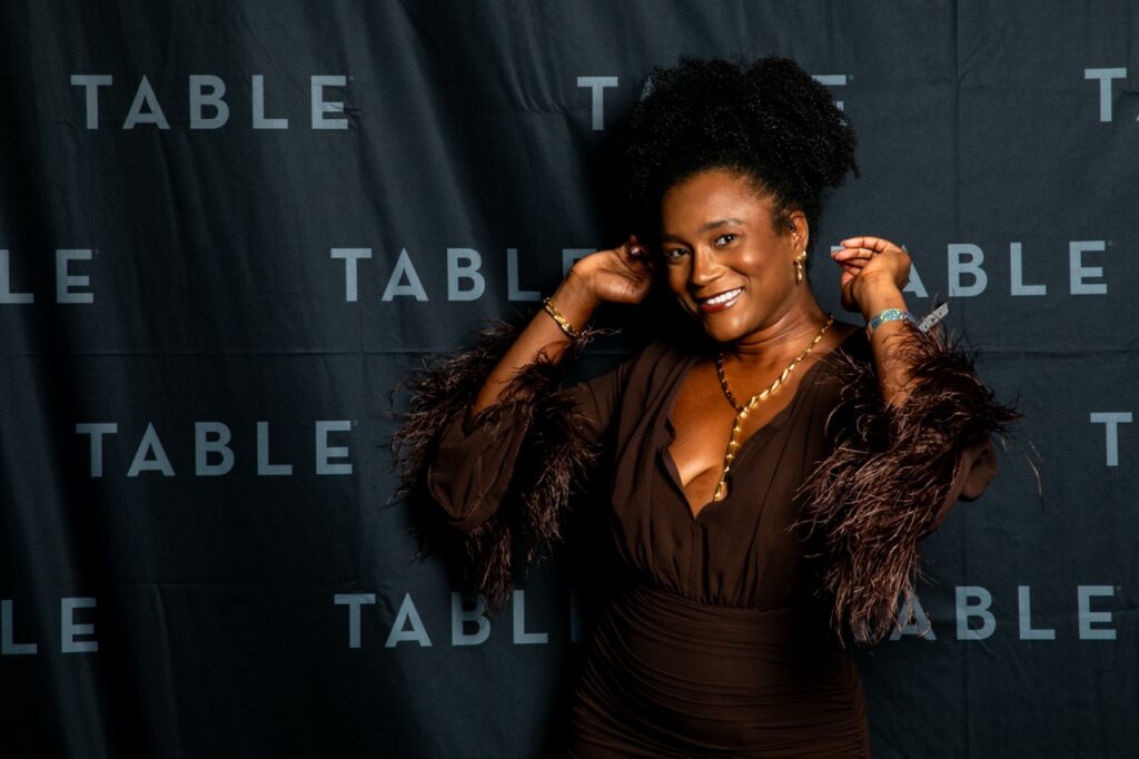 Woman in a brown dress with feathered sleeves posing in front of a black backdrop with "TABLE" repeated