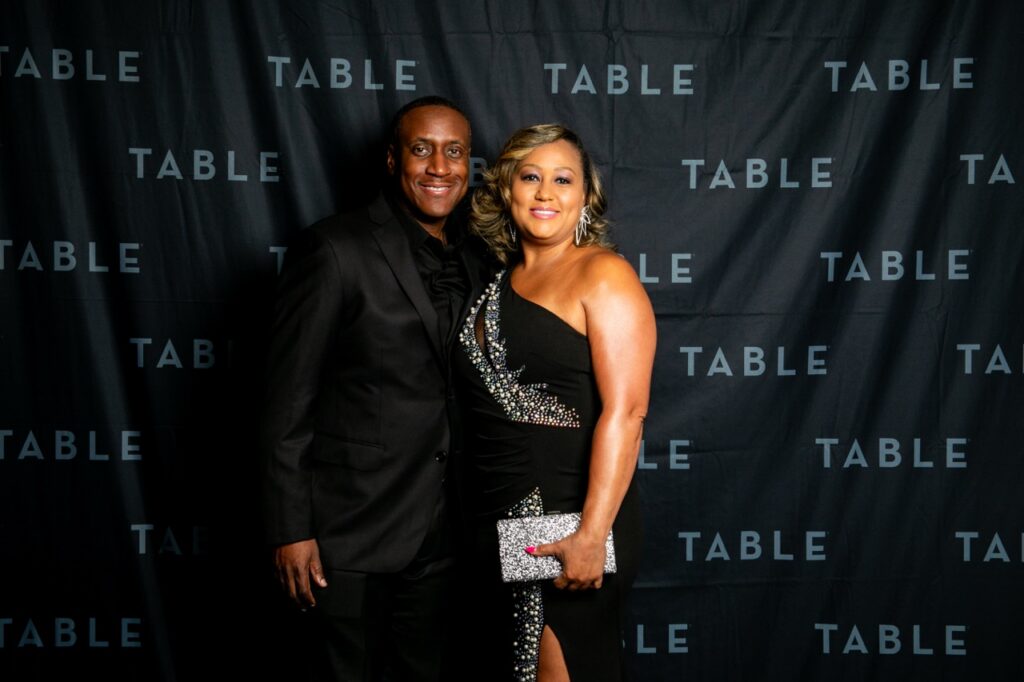 Couple in formal attire poses for a photo against a black backdrop with "TABLE" logo.