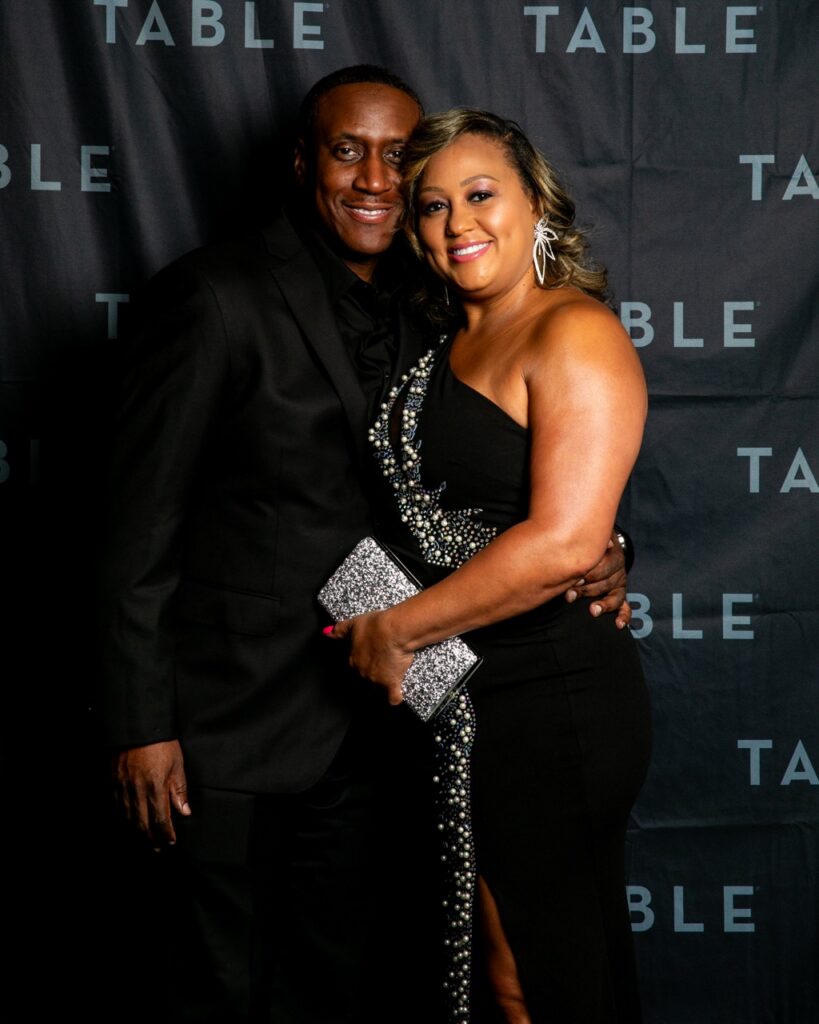 Couple in formal attire at an event with "TABLE" backdrop.