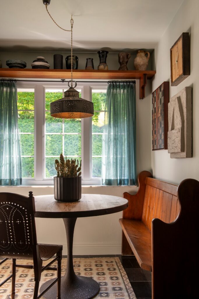 Breakfast nook with round wood pedestal table, wooden bench, sheer teal curtains, and ceramic pottery shelf.