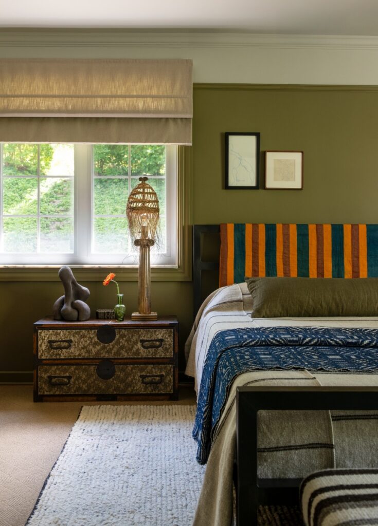 Olive green bedroom with striped headboard, blue patterned throw, and tassel lampshade on nightstand.