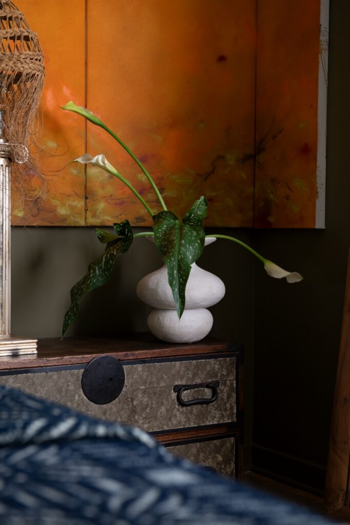 White textured ceramic vase with calla lilies on dark wood nightstand next to orange abstract art.