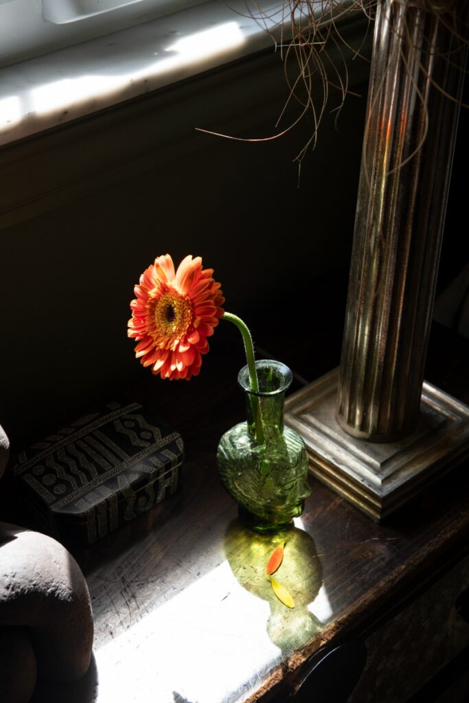 Sunlit nightstand detail with green glass face vase, orange gerbera daisy, and column lamp base.