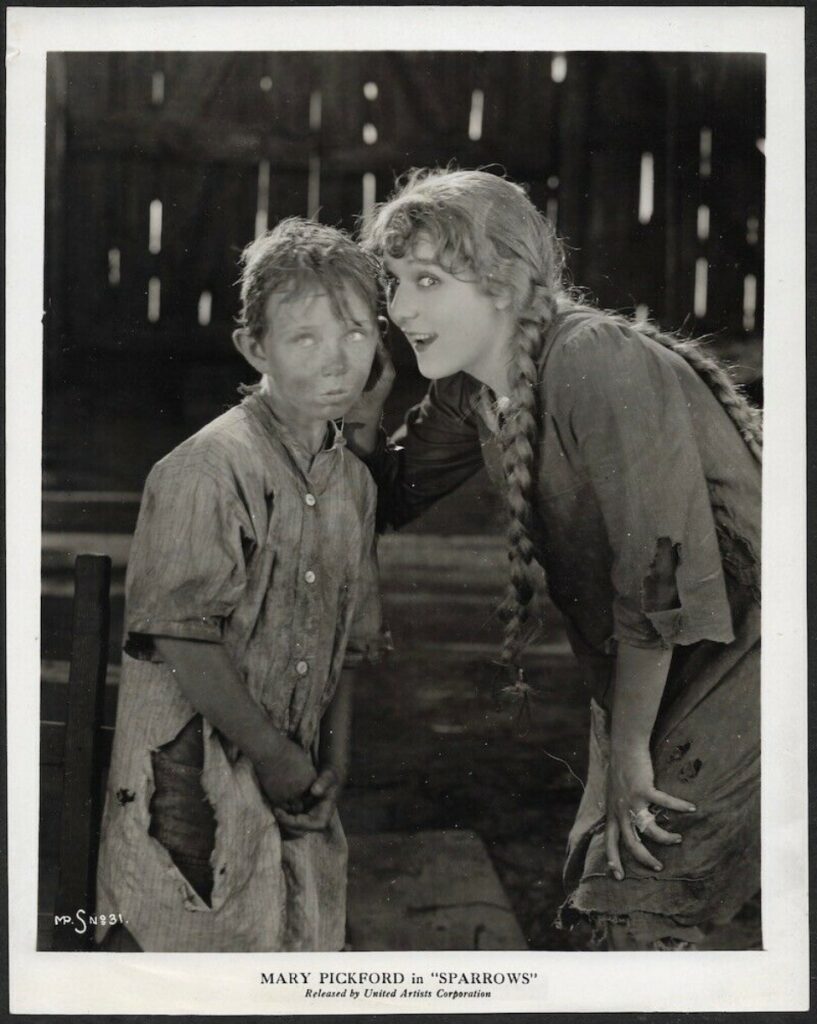 Black-and-white promotional still from Sparrows showing actress Mary Pickford with braided hair leaning in to whisper into the ear of a young boy in ragged clothing.
