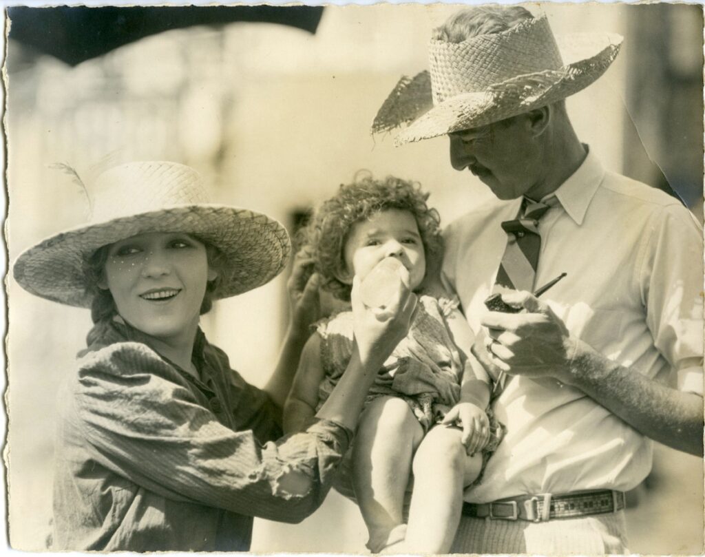 Vintage behind-the-scenes photograph featuring Mary Pickford in a straw hat smiling next to a man holding a toddler and a pipe.
