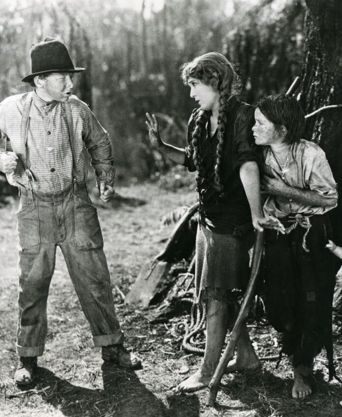 Black-and-white film still from Sparrows showing Mary Pickford protectively shielding a child as an older boy in suspenders and a hat gesticulates in front of them outdoors.