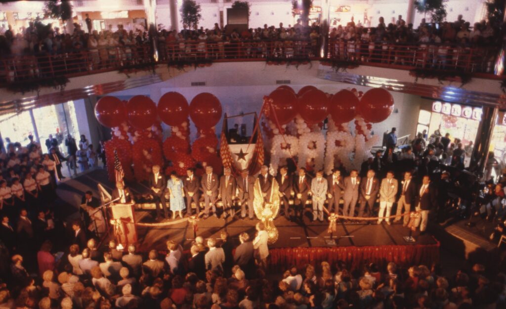 Crowd watches ribbon cutting ceremony with balloons and banners in a mall