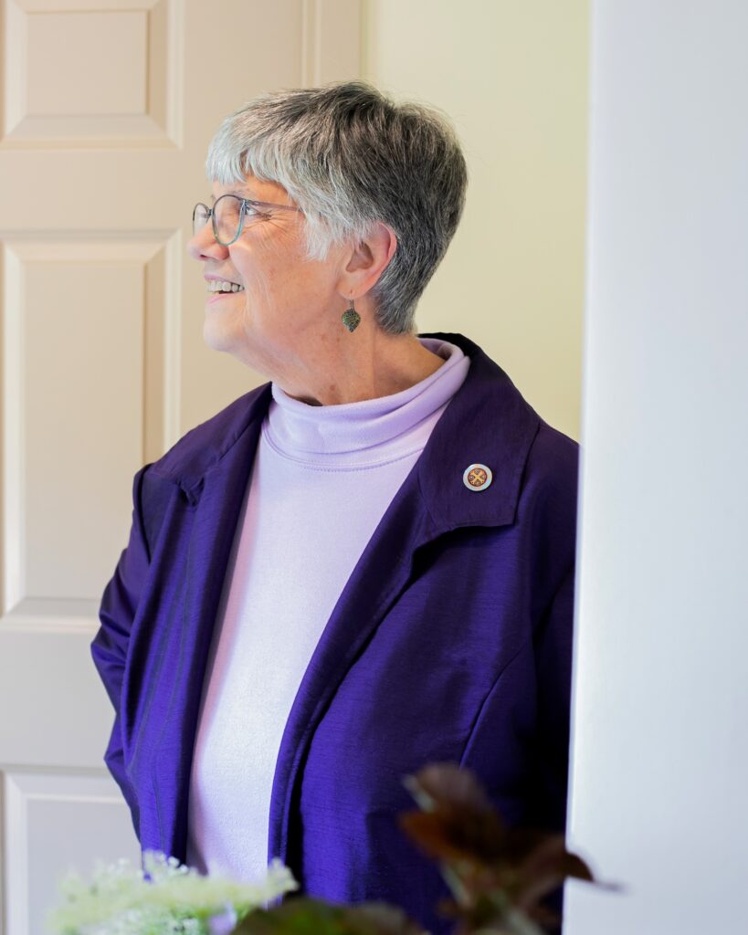 Elderly woman with gray hair and glasses wearing a purple jacket and a light purple turtleneck.