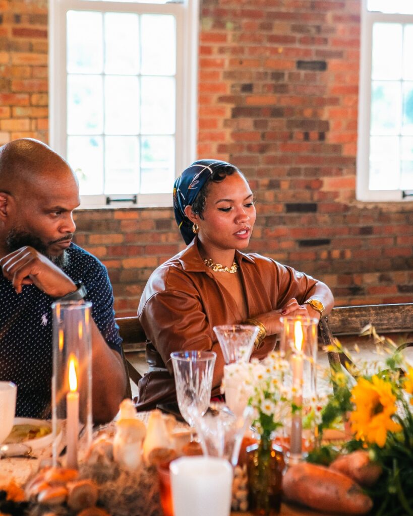 A woman in a brown leather shirt and headscarf listening attentively at the dinner table.