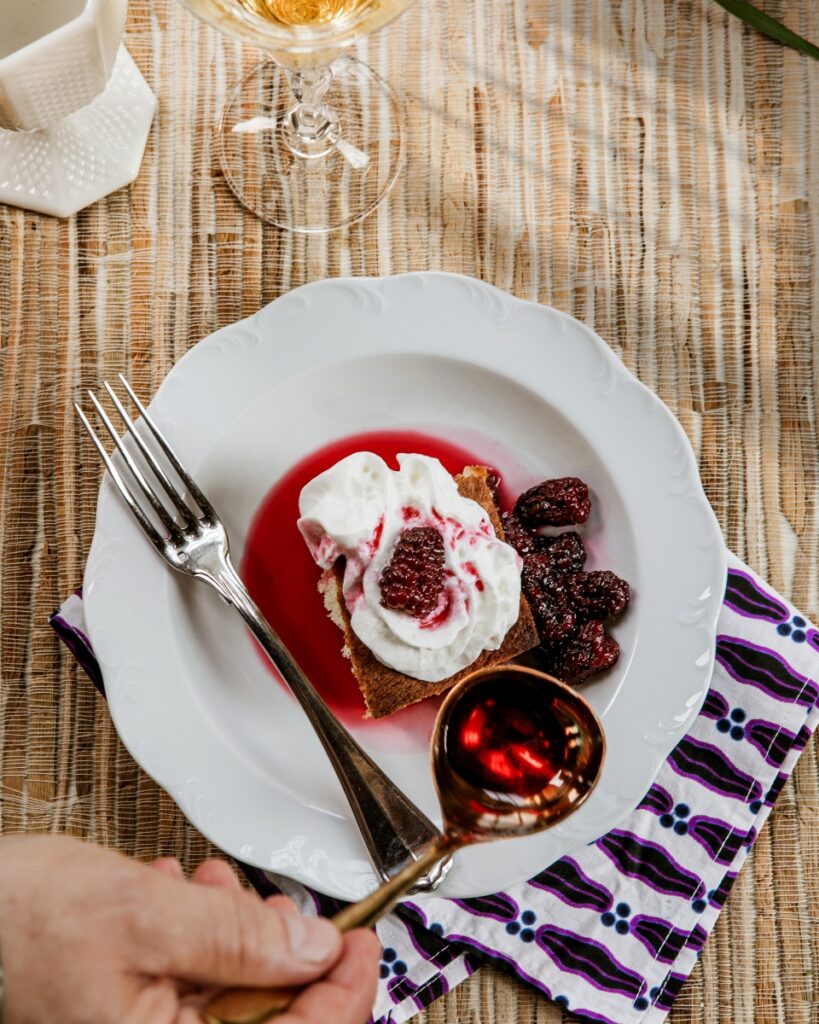 Square slice of Busy Day Cake topped with whipped cream, stewed blackberries, and berry syrup in a white bowl.