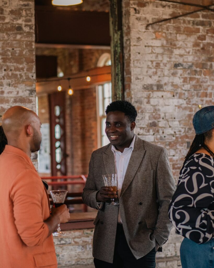 ony Ton-Aime wearing a gray suit jacket and holding a cocktail while mingling with event guests.