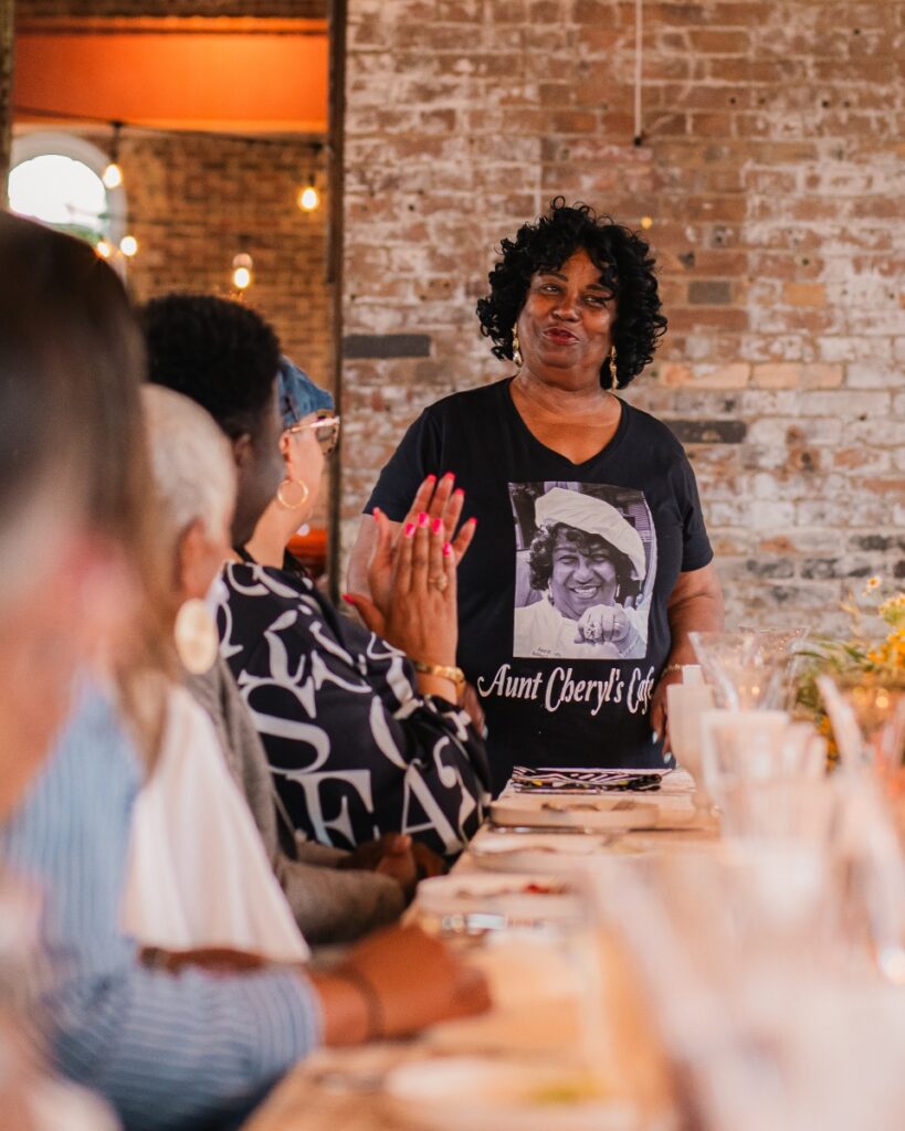 Cheryl Vance speaking to guests while wearing an Aunt Cheryl’s Café graphic t-shirt at the event table.