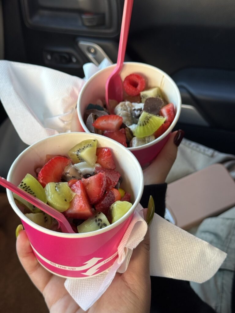 Two pink cups of frozen yogurt with fruit toppings, held by hands.