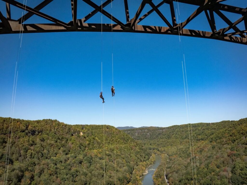 Two people rappelling from a bridge over a lush green valley.