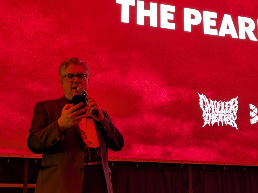 Man in suit speaks at Chiller Theater event with "The Pear" on screen