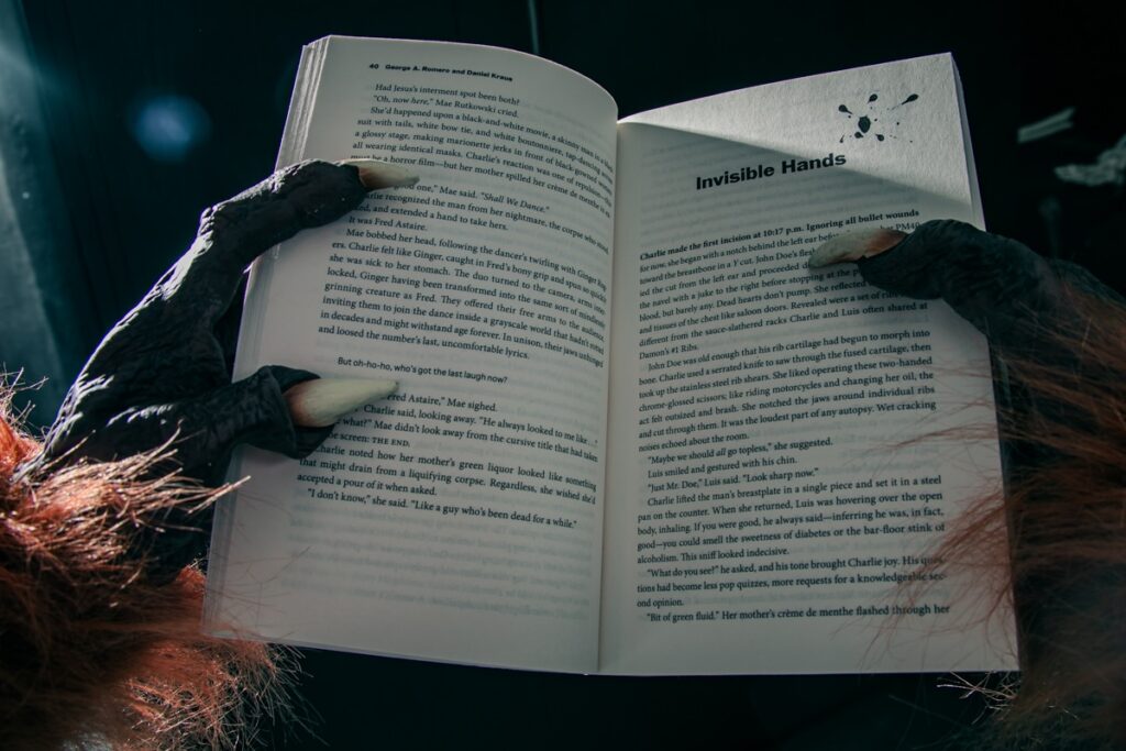 Monster hands turning pages of "Invisible Hands" book, hinting at George A. Romero's horror legacy.