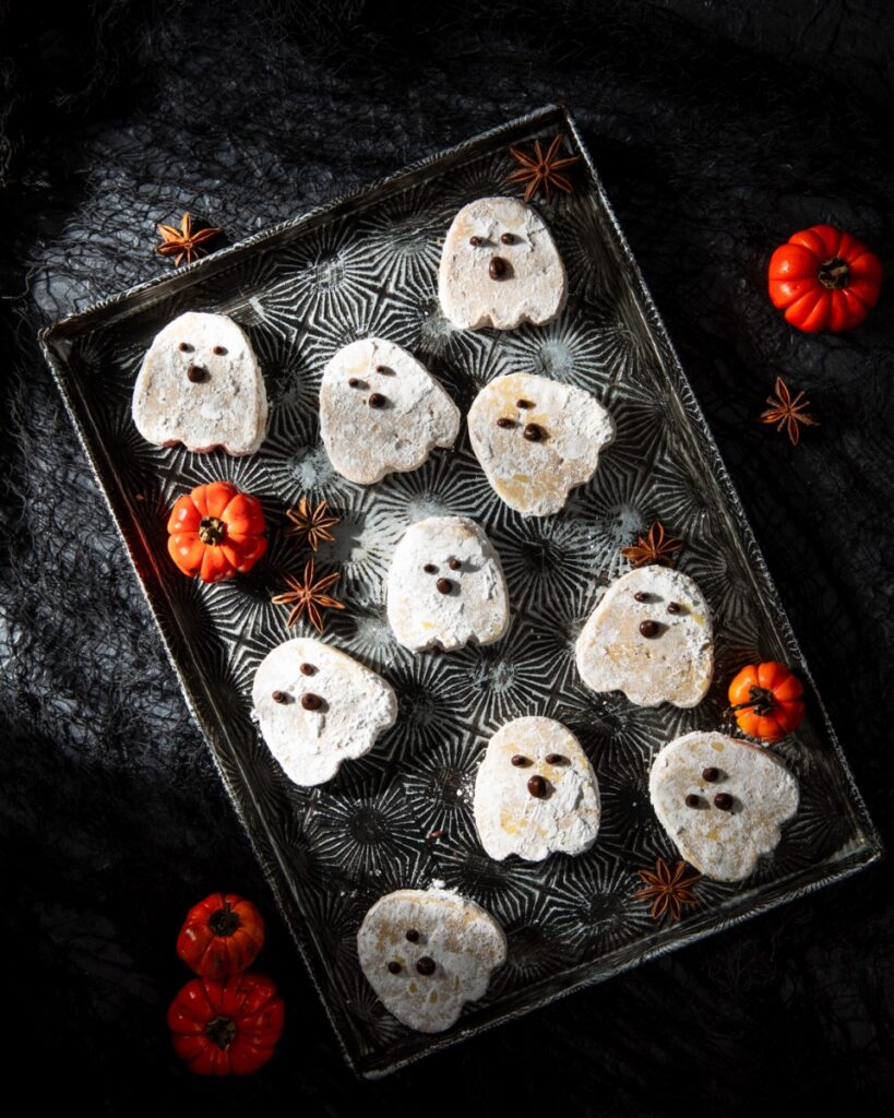 Ghost-shaped pumpkin brown butter shortbread cookies dusted with powdered sugar and decorated with melted chocolate faces, arranged on a vintage tray with mini pumpkins and star anise.