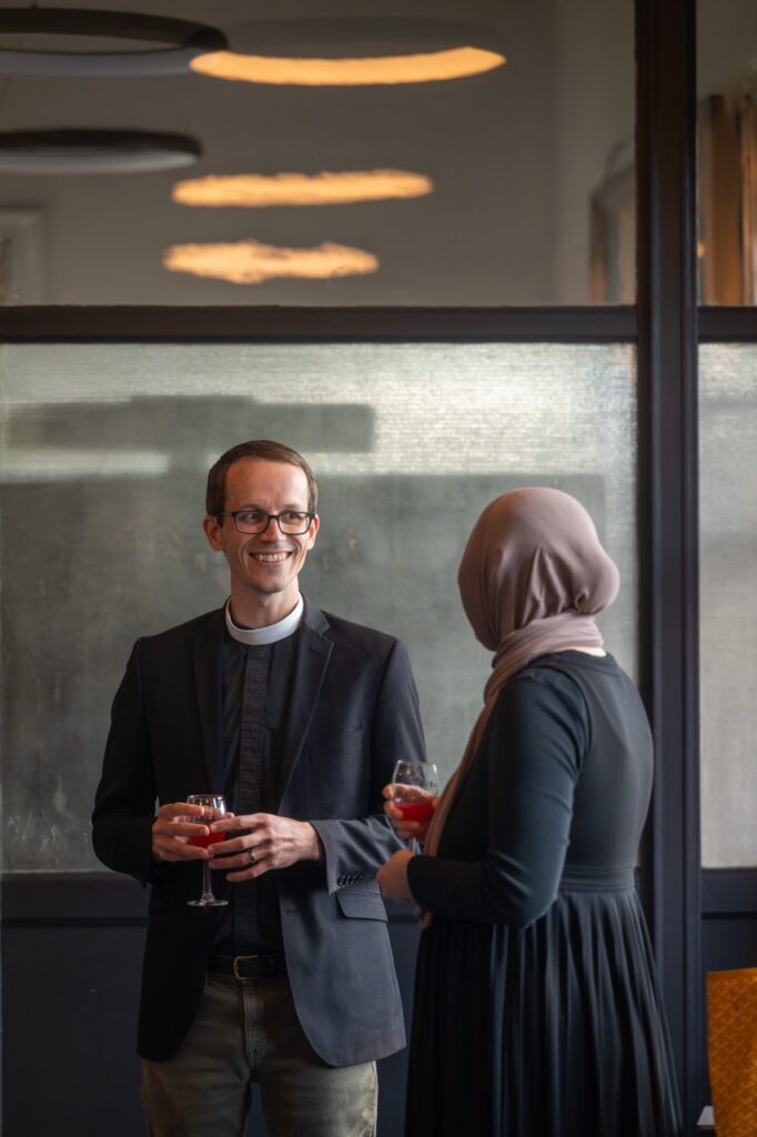 Reverend and woman in hijab holding drinks, discussing Pittsburgh's welcoming initiatives.